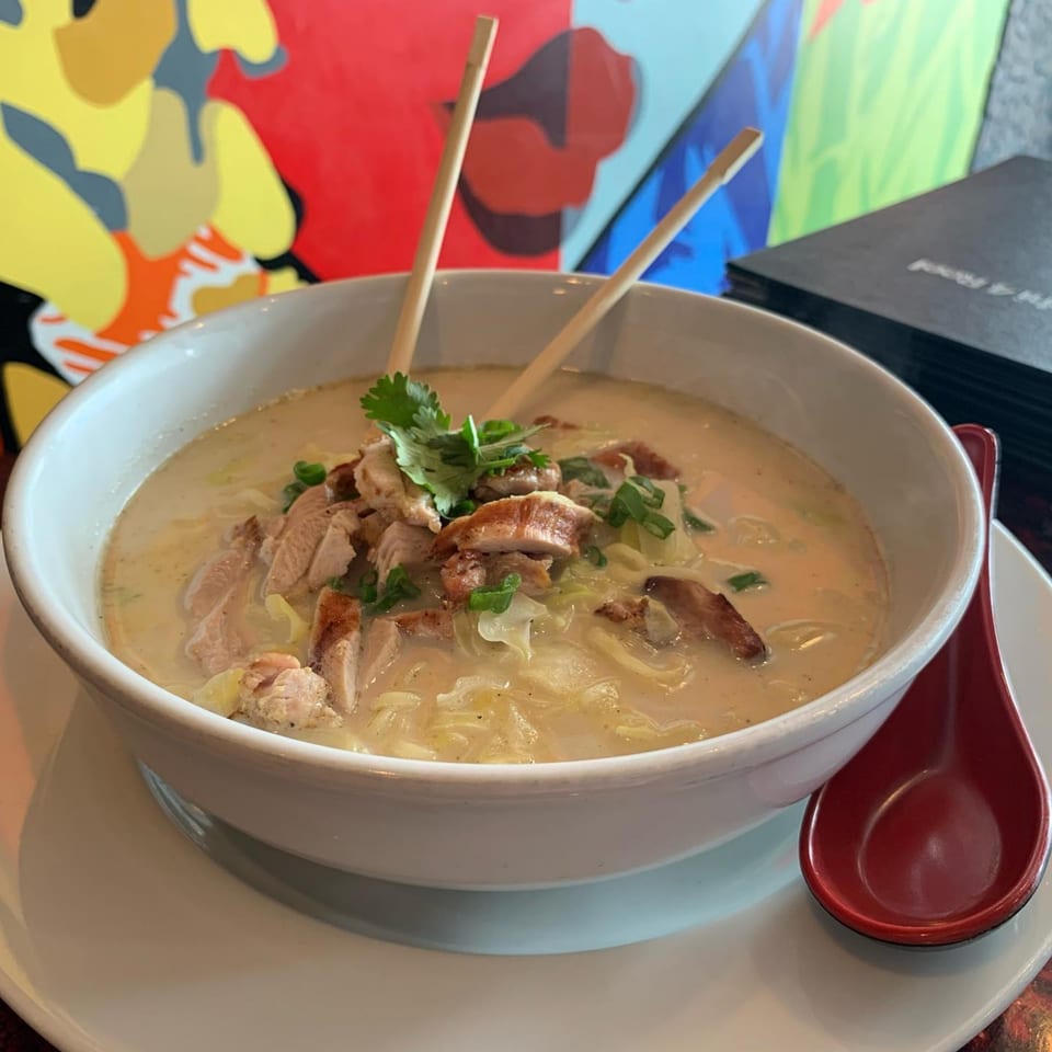 Creamy chicken ramen with chopsticks and a red spoon, against a colorful abstract background.